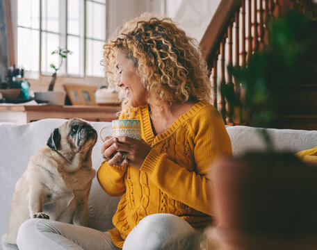 Woman with curly hair smiling on a sofa, holding a coffee mug while cuddling her elderly pug, sharing a cozy, joyful morning of quiet companionship at home