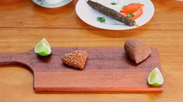 Hands serving traditional Lebanese and Syrian kibbeh and kofta on wooden table with lime wedges, authentic Middle Eastern cuisine concept featuring fried bulgur croquettes and grilled meat dishes