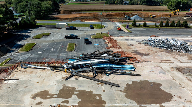 Workers clear debris from a demolished building in a rural area. Piles of bricks and scattered materials fill the lot. Construction vehicles and equipment are visible in the background.
