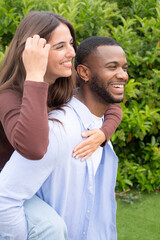 Fototapeta premium Happy interracial couple enjoying a playful piggyback in a green garden, smiling and affectionate together