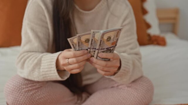 Woman counting dollars with hands while sitting crosslegged on a bedroom bed in pajamas; savings comfort.