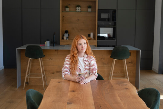 Young redhead woman sitting at wooden kitchen table looking thoughtful while holding smartphone in modern home