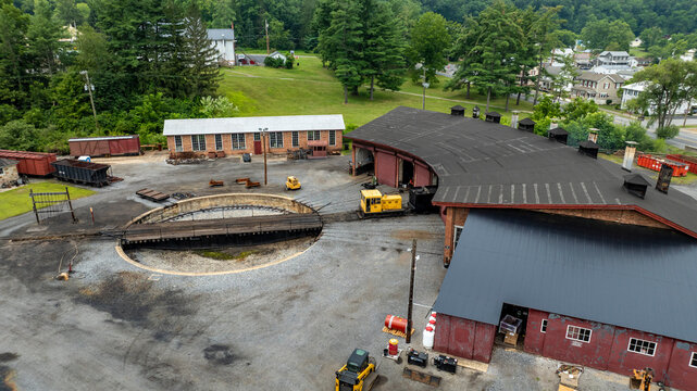 Workers manage train cars and maintenance activities at a roundhouse. The scene features a circular track and various tools around. Green trees and buildings are visible in the background.