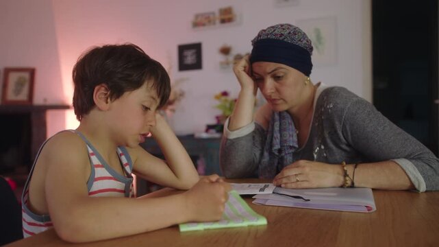 Mother undergoing chemotherapy helping child with homework at home table, headscarf parent supporting schoolwork with focused attention, everyday parenting and resilience