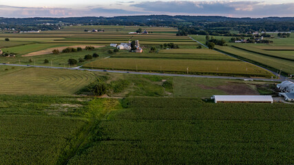Naklejka premium A vast area of farmland shows green fields stretching out under a colorful sky at sunset. A road runs through the landscape with buildings scattered nearby, highlighting rural life.