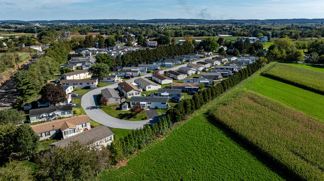 A view shows a cluster of mobile, manufactured homes aligned in rows arranged along a curved road. Nearby, fields stretch out with crops. The scene captures a rural setting in clear weather.