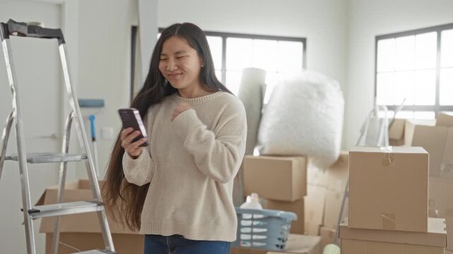 Woman holding a smartphone and smiling, fist pump gesture while standing among cardboard moving boxes, ladder and packing materials in a building; new home joy.
