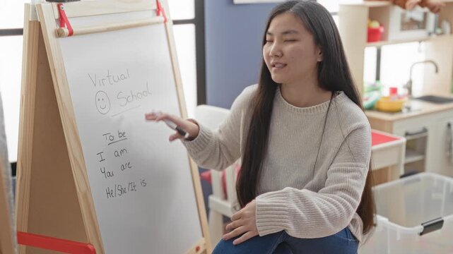 Woman teacher gestures with marker at whiteboard in studio, pointing hand visible while explaining present tense verbs to camera; engaged learning connection.