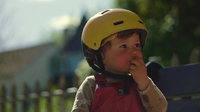 Toddler wearing yellow helmet sitting on bench outdoors, calmly eating tangerine piece in sunlight, candid everyday moment of childhood curiosity and simple snack time