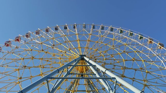 Huge ferris wheel with colorful cabins rotating at an amusement park. Low angle view of the spinning ride against a bright, cloudless sky
