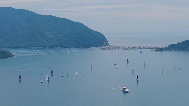 Aerial view of sailboats and yachts sailing across coastal bay with mountainous shoreline.