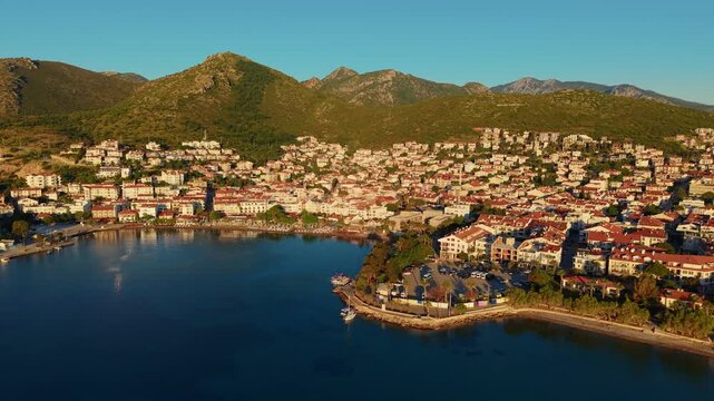 Aerial view of coastal town with red roof houses lining bay and mountain backdrop at sunrise, marina of Datcha Turkey.