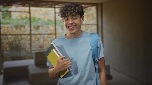 Teenage student male wearing a blue tee and backpack holds stacked notebooks to his chest while smiling and giving thumbs up inside a school building; optimism.