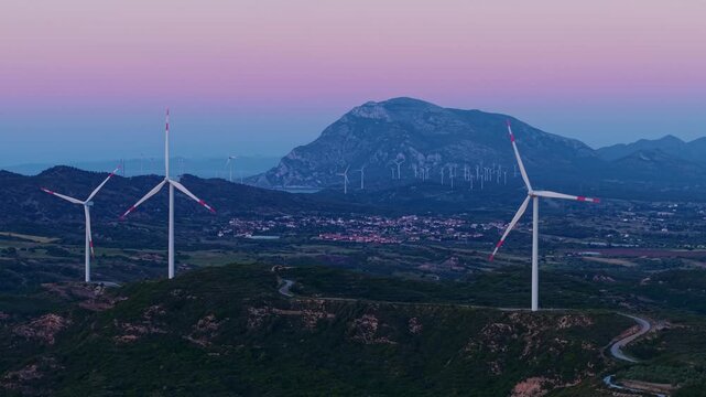 Aerial view of wind turbines rotating over rural valley and foothills at dusk with sunset.