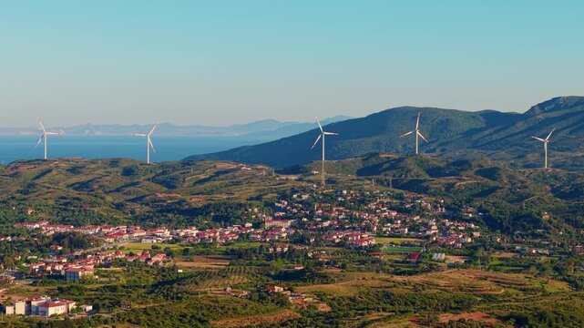 Aerial panning shot of wind turbines rotating above coastal village and terraced hills with sea horizon Datcha Turkey.