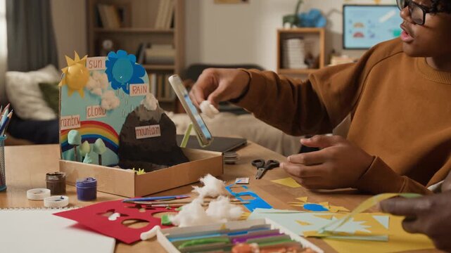 Medium close-up of devoted black father and son making creative science project on Earth, atmosphere, weather, natural cycles for school competition from color paper, paint, plasticine, cotton wool