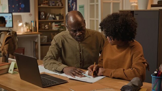 Medium shot of committed African American father explaining teenage son difficult mathematics problem, pointing to copybook and online content on laptop, both smiling, representing parenting, devotion