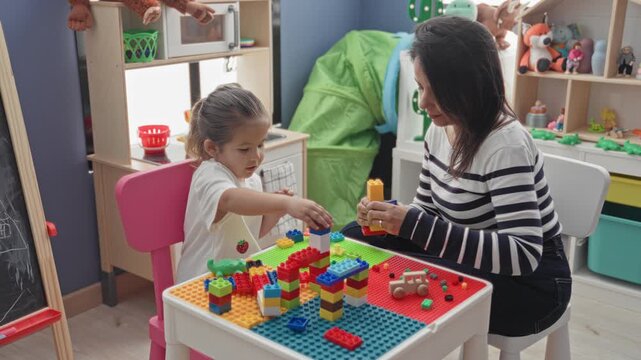 Woman helps little girl stack colorful building blocks with hands on table in playroom; bonding learning play.