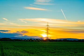 Obraz premium High-voltage transmission towers silhouetted against sunset sky, power lines crossing rural field, Sustainable energy infrastructure, green crops, distant hills, industrial infrastructure meets nature