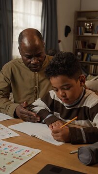 Medium vertical shot of supportive, smiling black father supervising little son learning to write by hand in copybook, praising, encouraging, while doing elementary school homework to achieve literacy