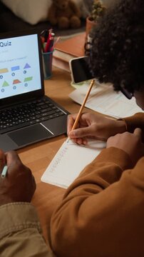 Vertical over-shoulder shot of young student solving geometry problem or mathematics equation during homeschooling lesson with father or tutor, laptop with online lesson on desk