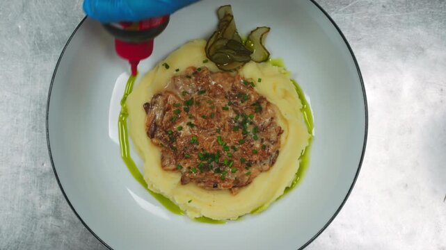 Top view of a chef hands in gloves plating a gourmet meal. They drizzle green herb oil from a squeeze bottle around mashed potatoes with beef stroganoff. Final touch on a restaurant dish