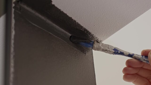 Close up of paintbrush applying dark paint along wall edge below ceiling