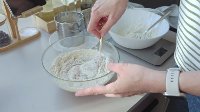 Hands mix flour in glass bowl with spoon beside white bowl on kitchen scale