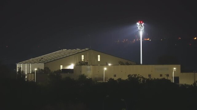 Static shot of an industrial building or warehouse at night. A powerful spotlight on a tall tower illuminates the area, creating a secure or mysterious atmosphere. Dark background with no people