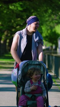Mother with headscarf pushing toddler in stroller along sunny park path, attentive parenting and daily family routine in nature, connection and calm outdoor lifestyle