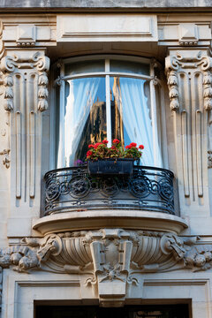 Quintessentially Parisian bow-window balcony adorned with vibrant red geraniums in a window box, framed by exquisite carved stone pilasters and a delicate curved wrought-iron railing. Avenue de