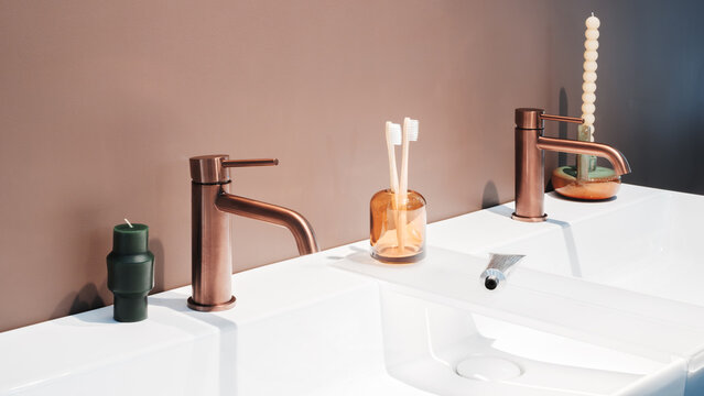 White countertop holds a modern sink. Copper faucet has a sleek handle. Two toothbrushes sit in a glass. Toothpaste tube rests beside them. Textured candle is in a ceramic holder