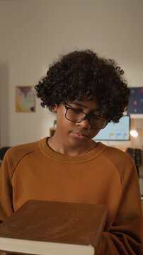 Medium vertical shot of 13-year-old middle school African American student in nerdy glasses blowing dust off neglected textbook while preparing to return to school for new academic year