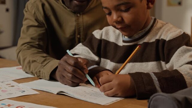 Tilt-down medium close-up of cheerful African American grandfather and grandson laughing during handwriting practice, pointing to copybook, joking about scribbles while preparing for elementary school