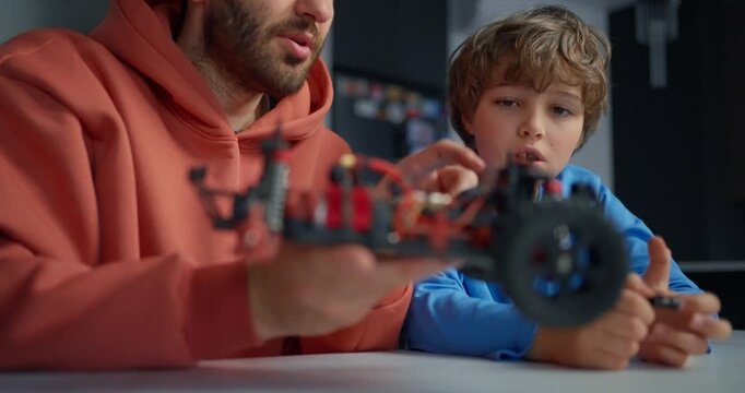 Father and son examining a remote control car chassis with exposed wires and wheels, sharing a hands-on robotics hobby project at a table