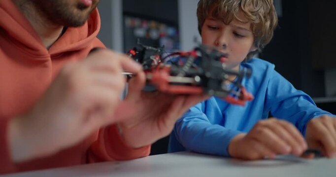 Father and young son examining a small homemade robotic vehicle at a table, learning mechanics and electronics together indoors