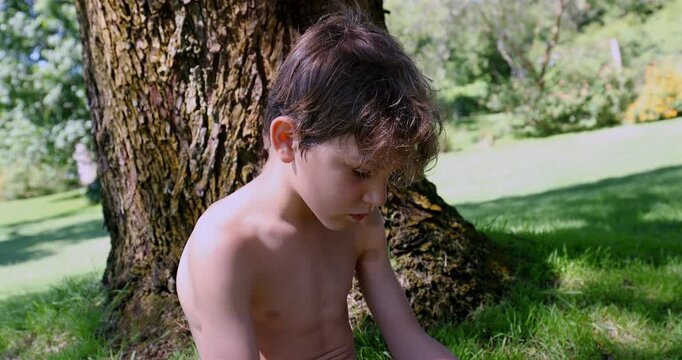 Boy seated on grass with legs extended, focused on smartphone held in hands, sitting near tree during quiet moment in summer park