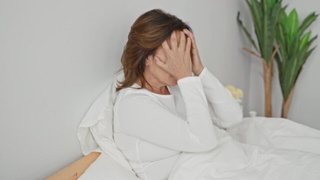 Woman in bed covering face then hands to head tying hair beside white duvet and plant in bedroom; morning routine fatigue.