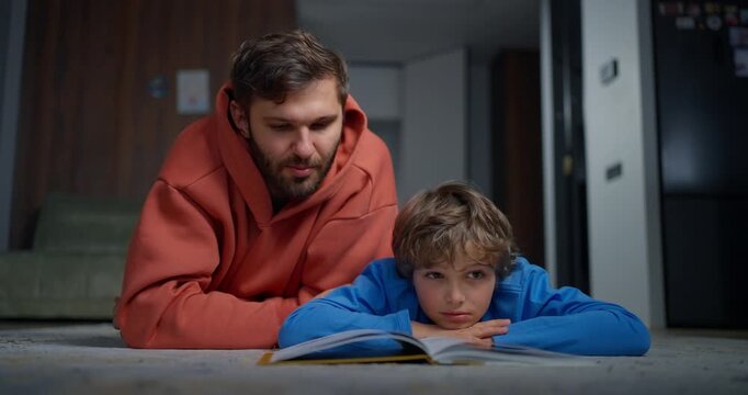 Caring father and young son lying on the living room floor reading a book together, sharing quiet homework time and family bonding at home