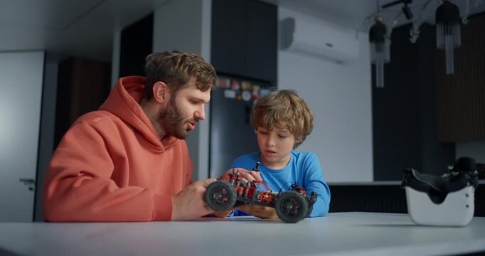 Father and young son work together assembling a remote controlled toy car at a kitchen table, sharing a focused hands on learning moment