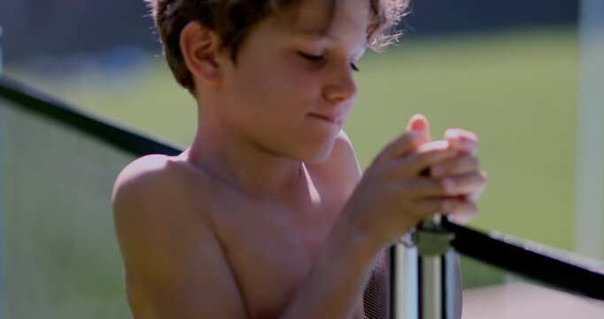 Closeup of shirtless boy&rsquo;s hands and face as he tries to unlock swimming pool gate, looking down at latch with focused expression on sunny day