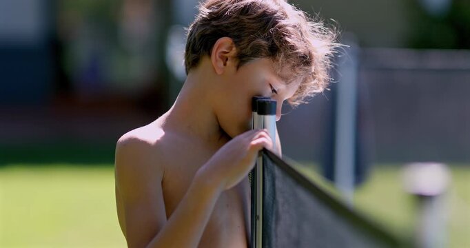 Shirtless boy resting head on swimming pool safety fence post during sunny day, eyes closed in peaceful moment by outdoor yard barrier near residential pool
