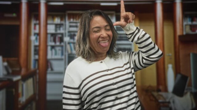 Woman sticks tongue out and makes l hand sign near forehead while smiling in a library building; playful mischief expression.