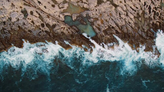 Waves crashing onto a textured rocky shoreline with tide pools from above