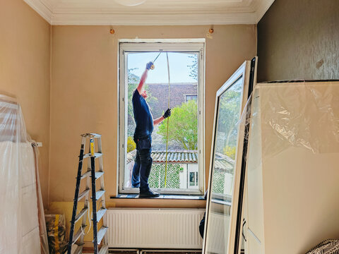 A builder measures a window opening to install a new frame in a bedroom.