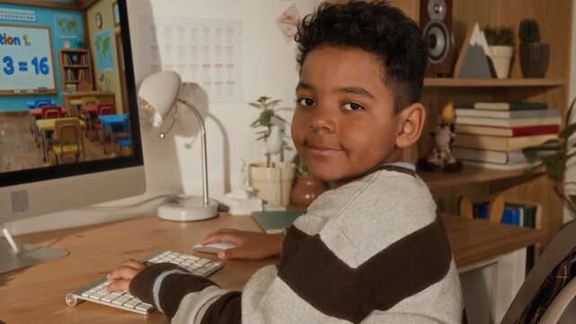 Medium portrait of confident 6-year-old African American boy sitting at home desk, using interactive e-learning app for doing math sums, turning to camera and smiling, preparing for elementary school