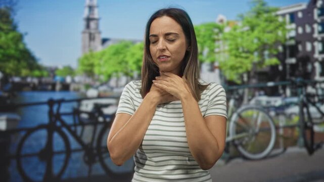 Woman young hispanic holding throat with both hands beside a parked bicycle on a street by a canal and bridge; breathing difficulty anxiety.