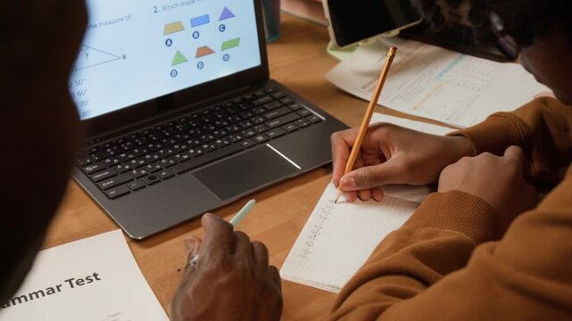 Over-shoulder of black schoolboy solving difficult geometry problem, writing equation in copybook with guidance from dedicated father, laptop with online learning content for homeschooling on desk