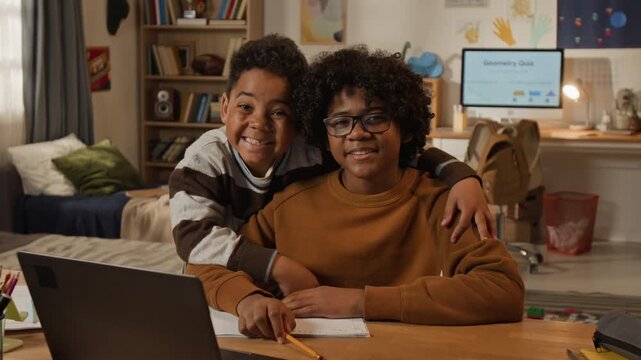 Medium portrait of two African American brothers posing in shared bedroom at home, teenage schoolboy studying on online edtech website on laptop, younger sibling hugging, looking at camera, smiling