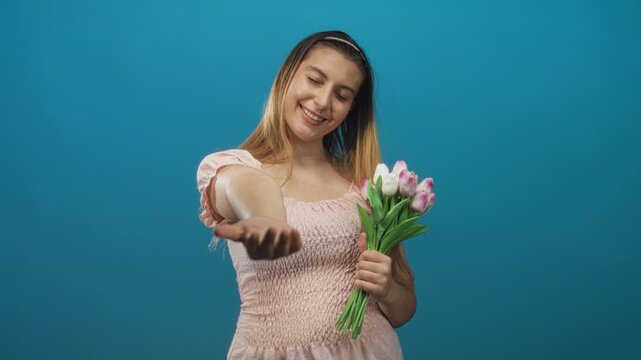 Woman in pink dress extends open hand offering pink and white tulip bouquet while smiling against a blue studio backdrop; giving warmth.